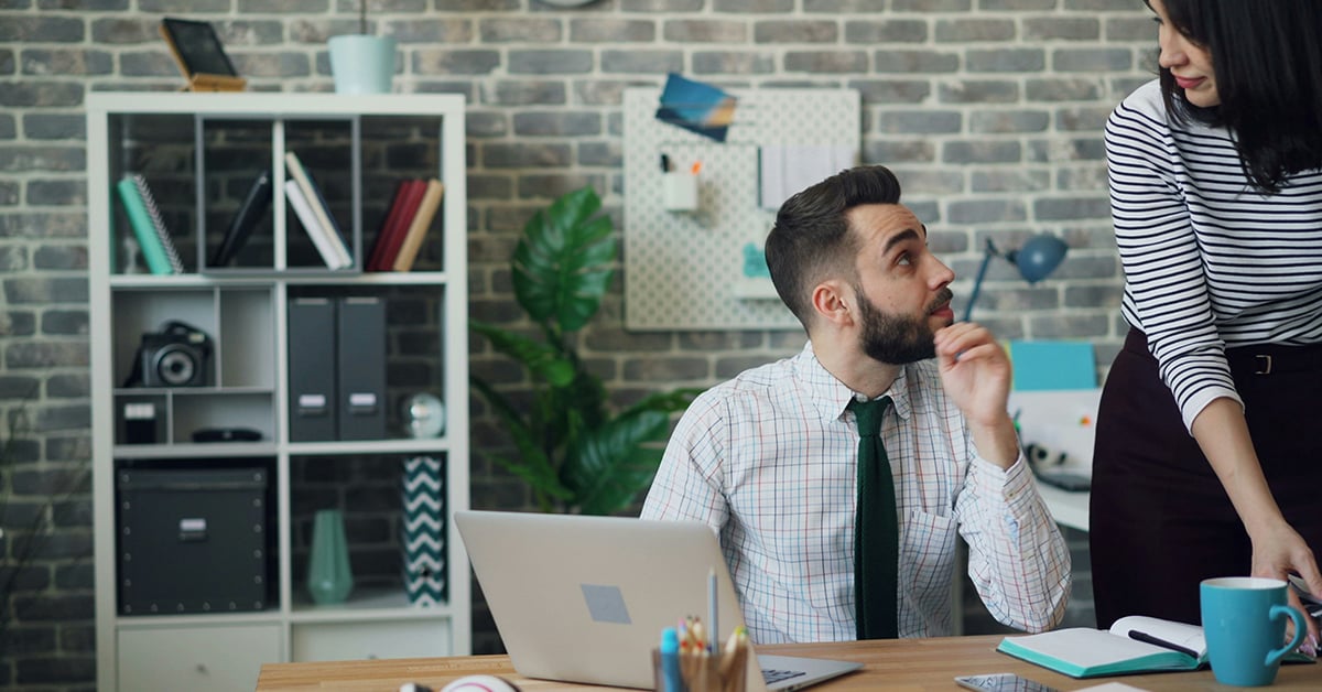 A man sits at a desk in an office, turning to his coworker to ask a question.